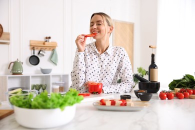 Photo of Smiling woman eating bell pepper while cooking at table in kitchen