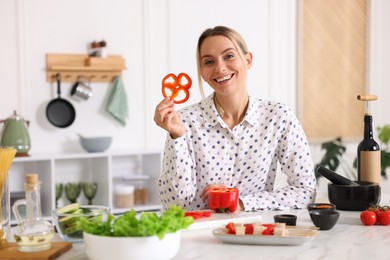 Photo of Smiling woman showing slice of bell pepper while cooking at table in kitchen
