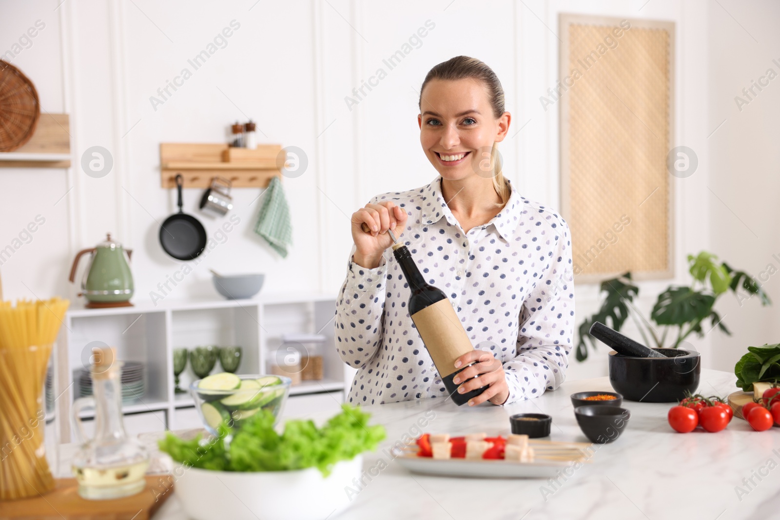 Photo of Smiling woman opening bottle of wine while cooking at table in kitchen