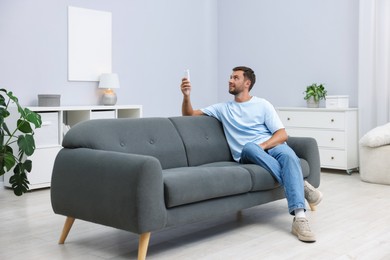 Photo of Man using remote to control air conditioner on sofa at home