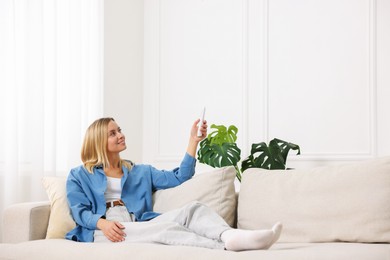 Photo of Woman using remote to control air conditioner on sofa at home