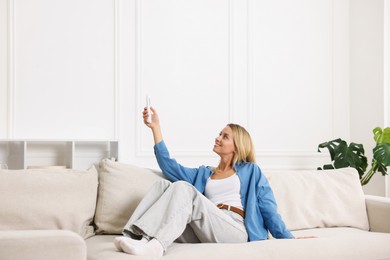 Photo of Woman using remote to control air conditioner on sofa at home