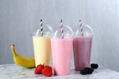 Photo of Milkshakes of different flavors in plastic cups and ingredients on white marble table, closeup