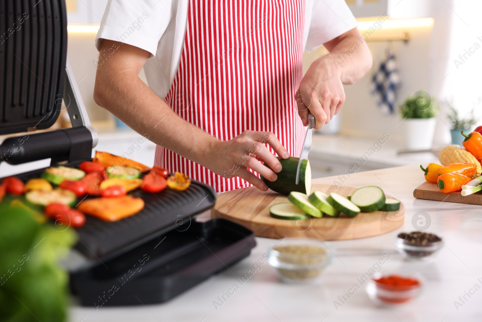 Photo of Man cutting zucchini while cooking vegetables on electric grill in kitchen, closeup