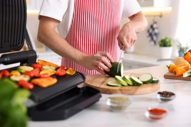 Photo of Man cutting zucchini while cooking vegetables on electric grill in kitchen, closeup