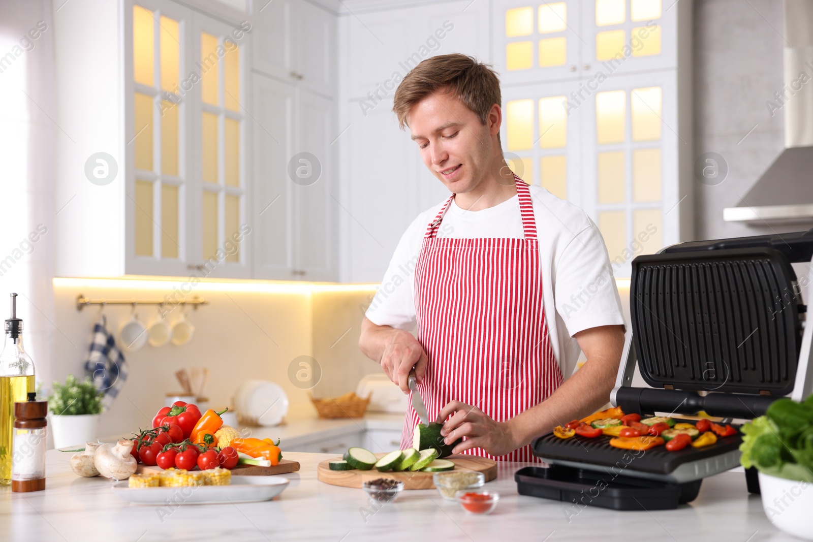 Photo of Man cutting zucchini while cooking vegetables on electric grill in kitchen