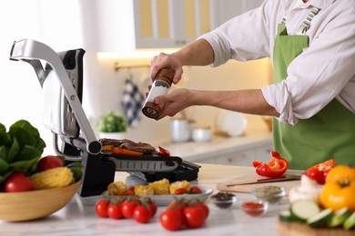 Photo of Man adding salt to vegetables and meat cooking on electric grill in kitchen, closeup