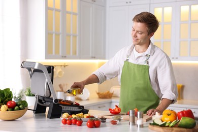 Photo of Man pouring oil onto vegetables cooking on electric grill in kitchen