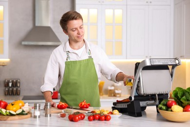 Photo of Man pouring oil onto vegetables cooking on electric grill in kitchen