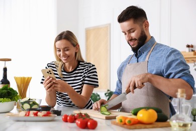 Photo of Cooking process. Smiling man cutting cucumber while his wife looking at recipe on smartphone at table in kitchen