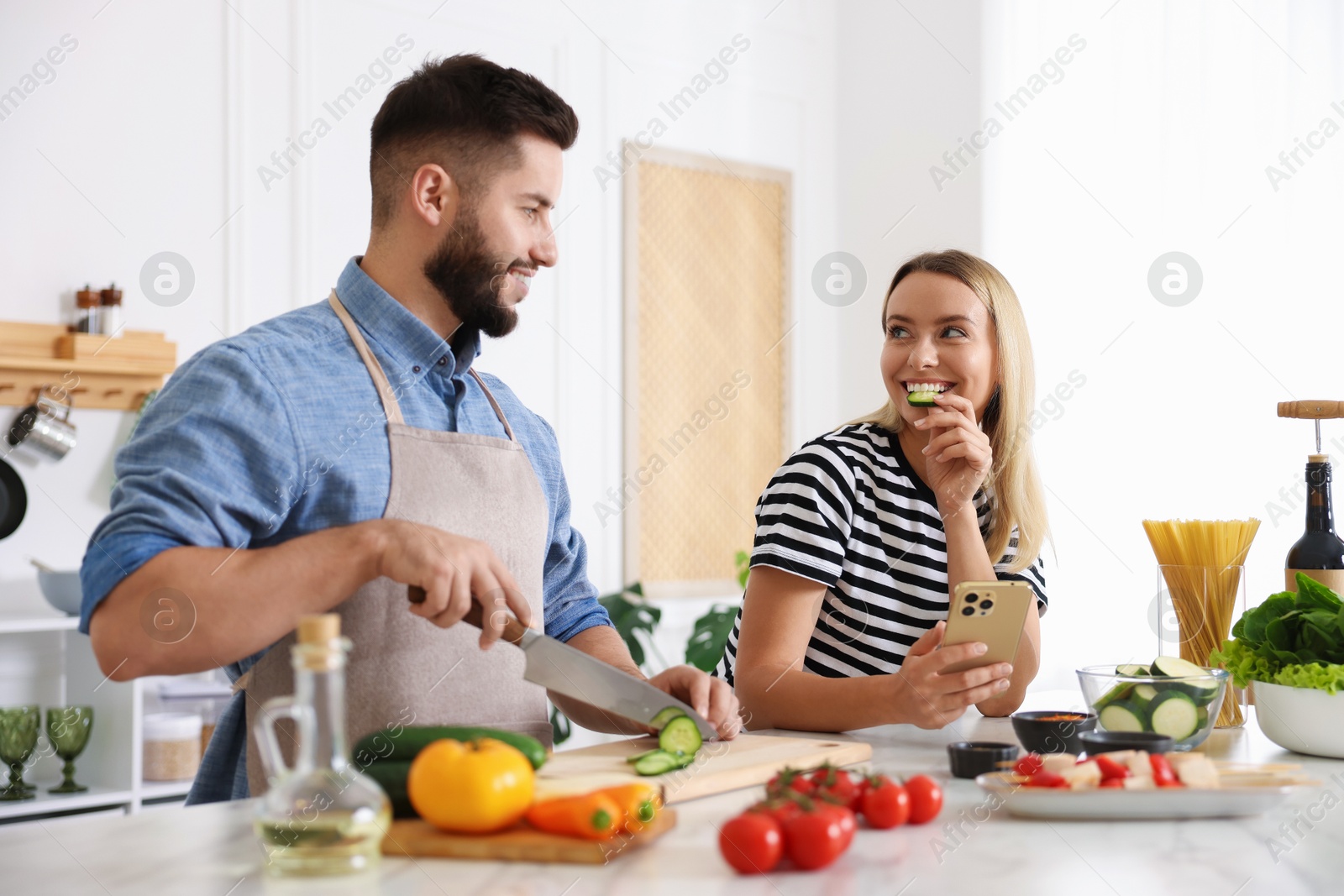 Photo of Cooking process. Smiling man cutting cucumber while his wife looking at recipe on smartphone at table in kitchen