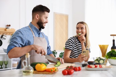 Photo of Cooking process. Smiling man cutting cucumber while his wife looking at recipe on smartphone at table in kitchen