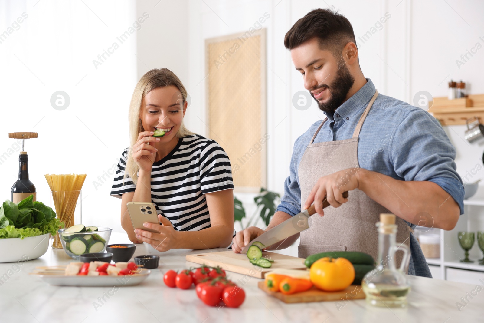 Photo of Cooking process. Smiling man cutting cucumber while his wife looking at recipe on smartphone at table in kitchen