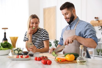 Photo of Cooking process. Smiling man cutting cucumber while his wife looking at recipe on smartphone at table in kitchen