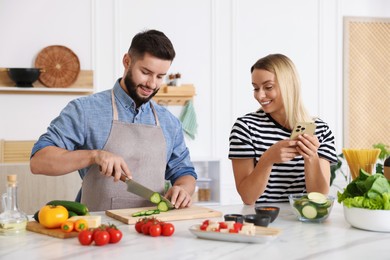 Photo of Cooking process. Smiling man cutting cucumber while his wife looking at recipe on smartphone at table in kitchen