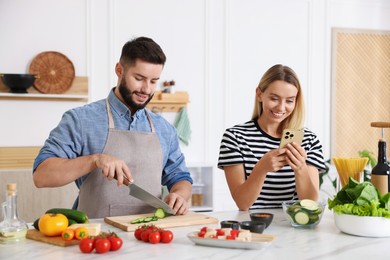 Photo of Cooking process. Smiling man cutting cucumber while his wife looking at recipe on smartphone at table in kitchen