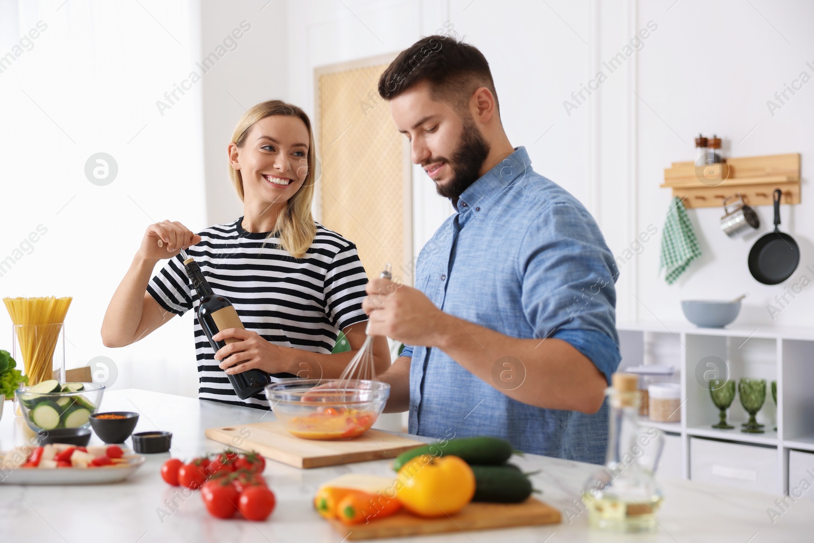 Photo of Smiling woman opening bottle of wine while cooking with her husband at table in kitchen