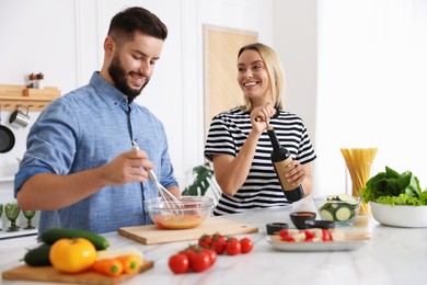 Photo of Smiling woman opening bottle of wine while cooking with her husband at table in kitchen