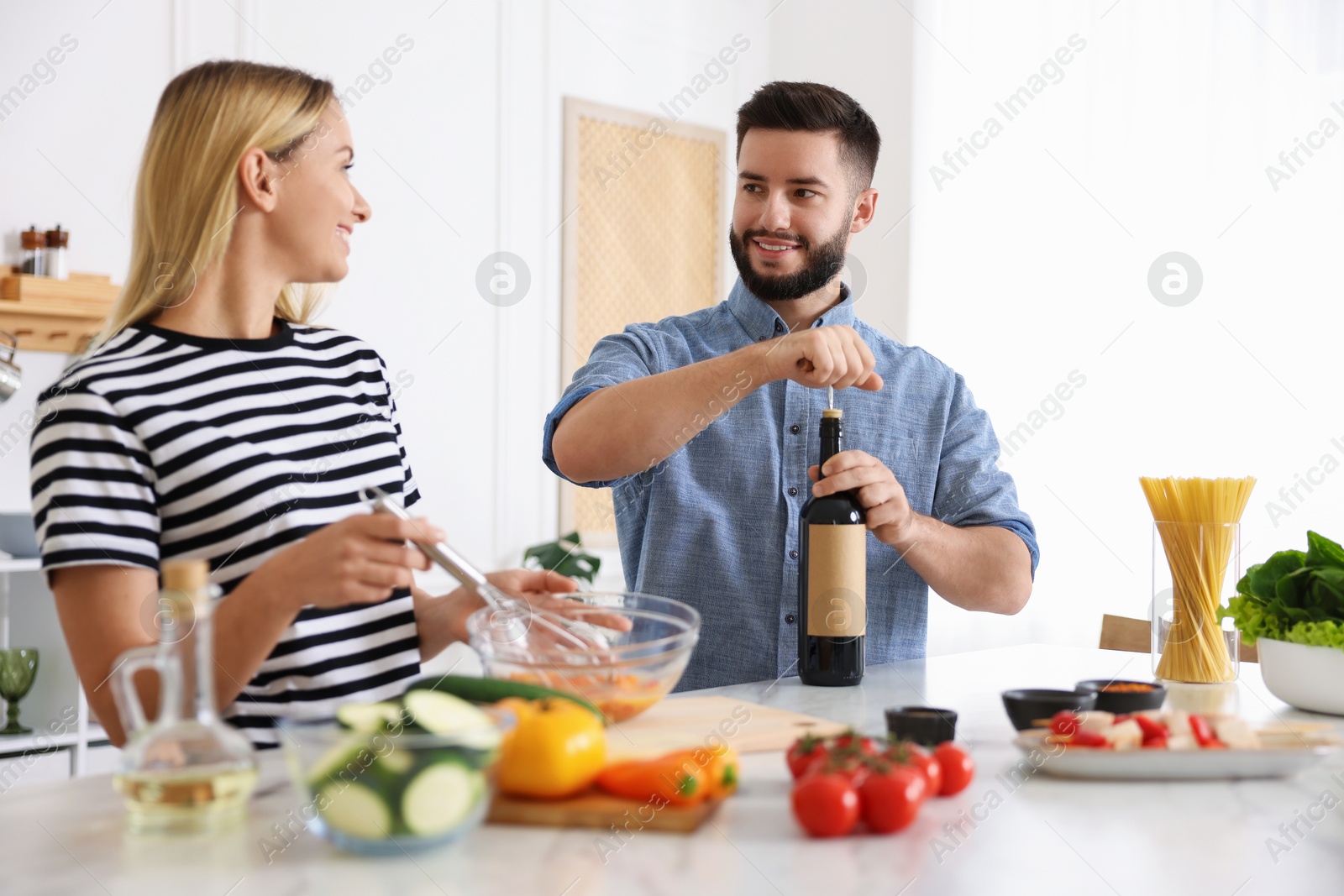 Photo of Smiling man opening bottle of wine while cooking with his wife at table in kitchen