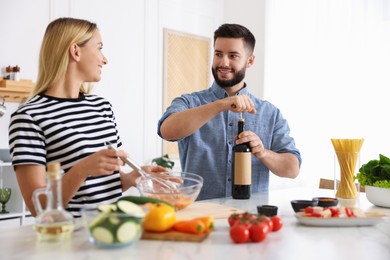 Photo of Smiling man opening bottle of wine while cooking with his wife at table in kitchen