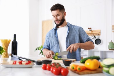 Photo of Smiling man looking at recipe on tablet while cooking at table in kitchen