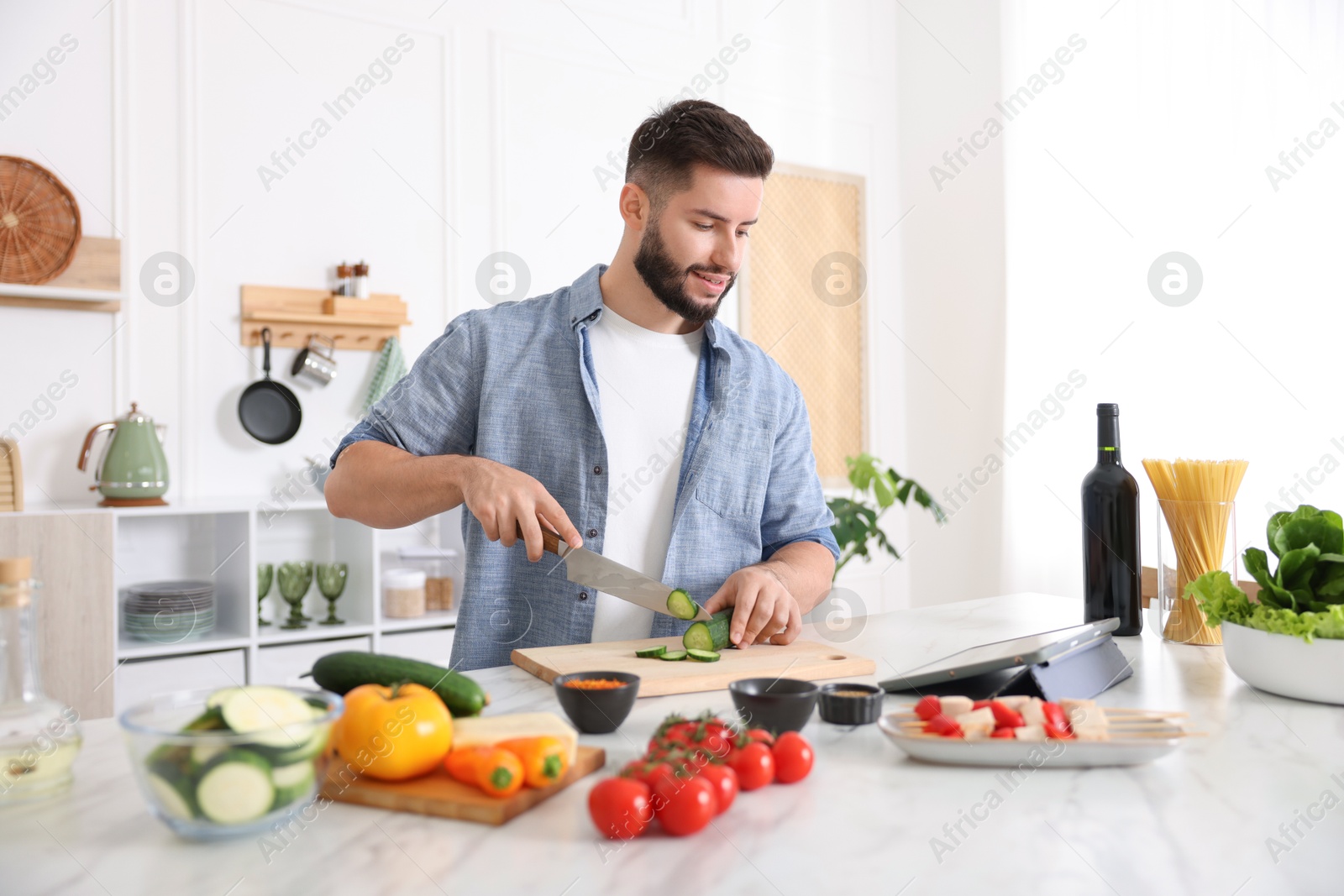 Photo of Smiling man looking at recipe on tablet while cooking at table in kitchen