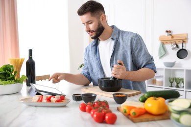 Photo of Young man looking at recipe on tablet while cooking at table in kitchen