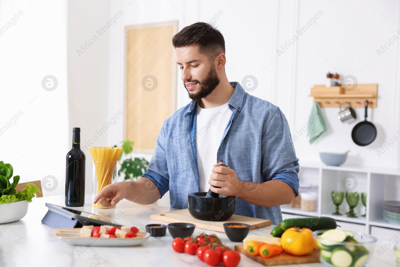 Photo of Smiling man looking at recipe on tablet while cooking at table in kitchen