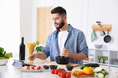 Photo of Smiling man looking at recipe on tablet while cooking at table in kitchen