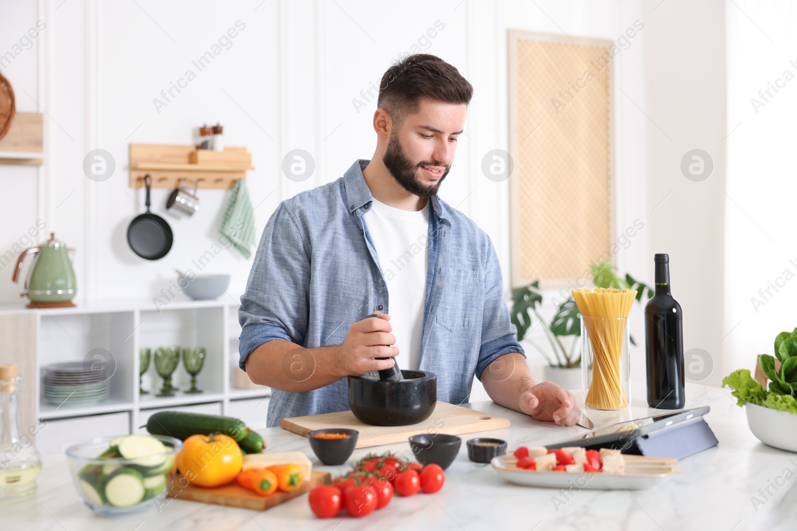 Photo of Smiling man looking at recipe on tablet while cooking at table in kitchen