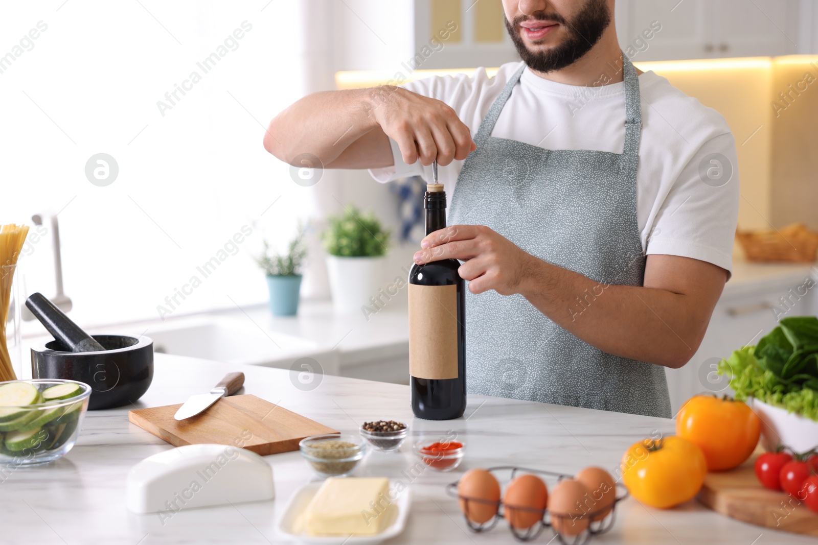 Photo of Man opening bottle of wine while cooking at table in kitchen, closeup