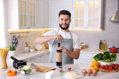 Photo of Smiling man opening bottle of wine while cooking at table in kitchen