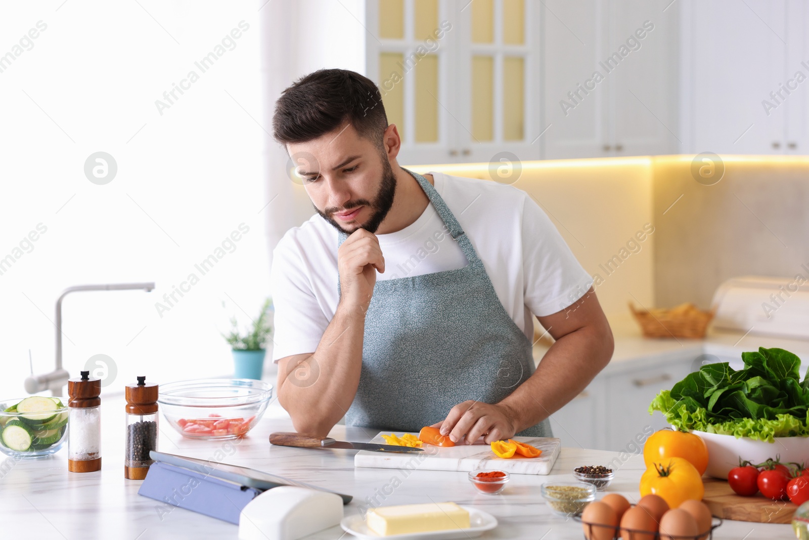 Photo of Young man looking at recipe on tablet while cooking at table in kitchen