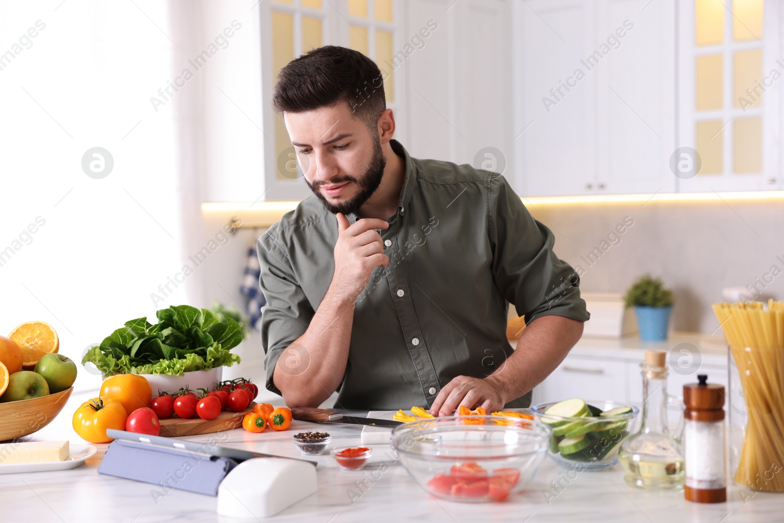 Photo of Young man looking at recipe on tablet while cooking at table in kitchen
