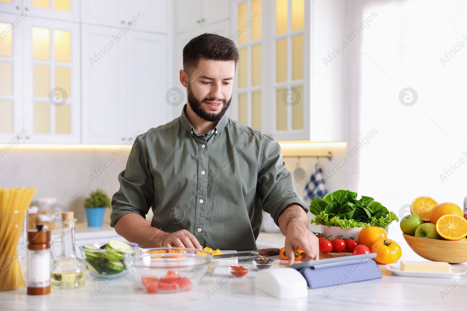 Photo of Smiling man looking at recipe on tablet while cooking at table in kitchen