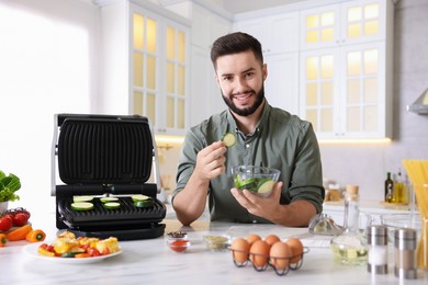 Photo of Smiling man cooking zucchini on electric grill at table in kitchen