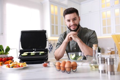 Photo of Smiling man cooking zucchini on electric grill at table in kitchen