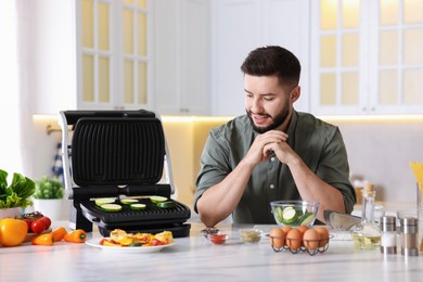 Photo of Smiling man cooking zucchini on electric grill at table in kitchen