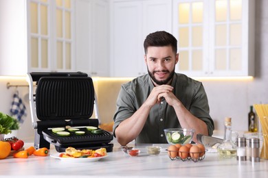 Photo of Smiling man cooking zucchini on electric grill at table in kitchen