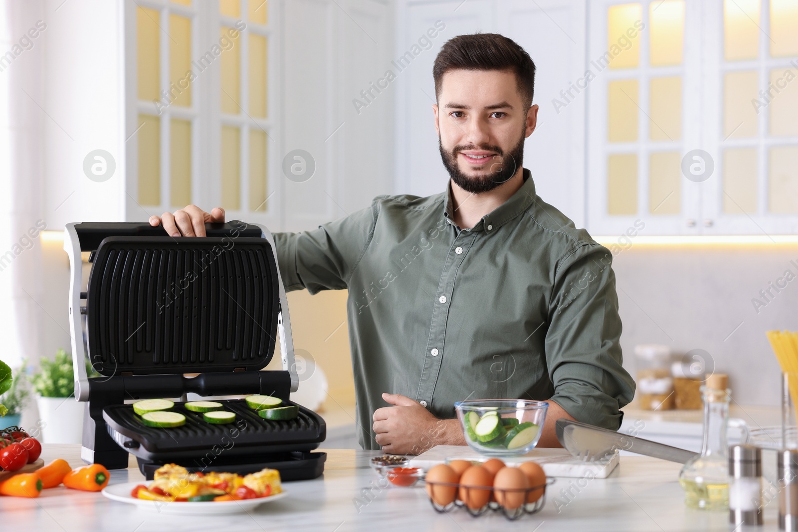 Photo of Smiling man cooking zucchini on electric grill at table in kitchen
