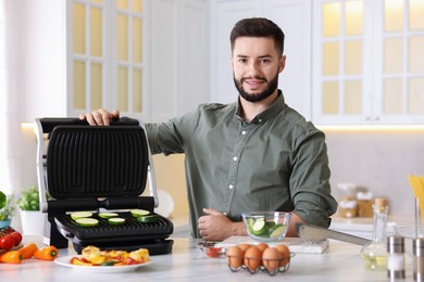 Photo of Smiling man cooking zucchini on electric grill at table in kitchen