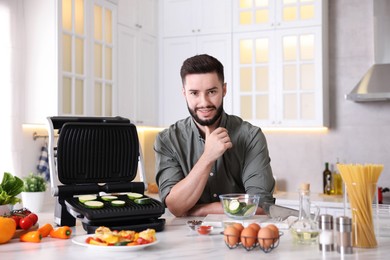 Photo of Smiling man cooking zucchini on electric grill at table in kitchen