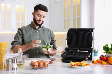 Photo of Cooking process. Young man with bowl of sliced zucchini near electric grill at table in kitchen