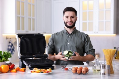 Photo of Cooking process. Smiling man with bowl of sliced zucchini near electric grill at table in kitchen