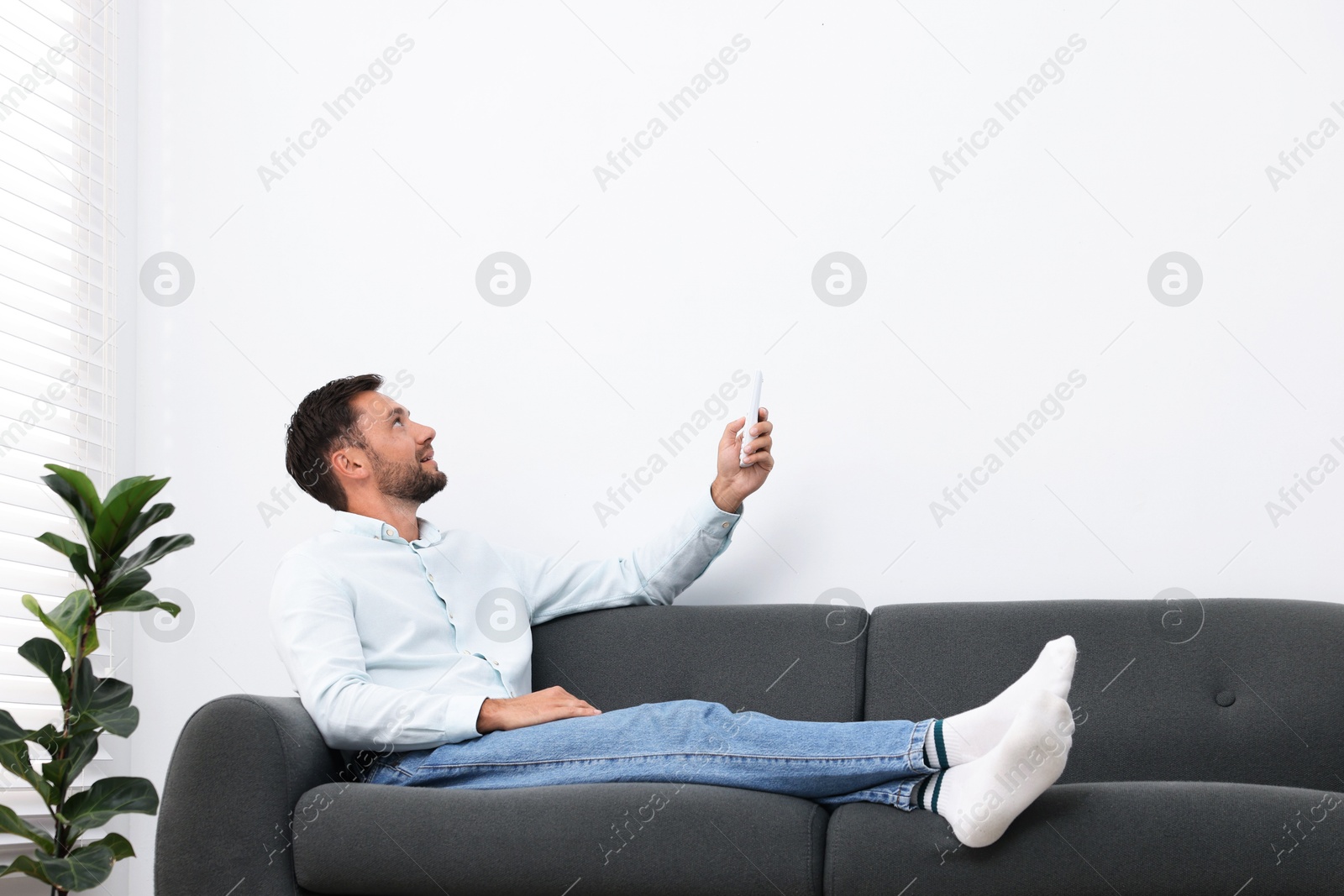 Man using remote to control air conditioner on sofa at home. Space for text Photo of Man using remote to control air conditioner on sofa at home. Space for text