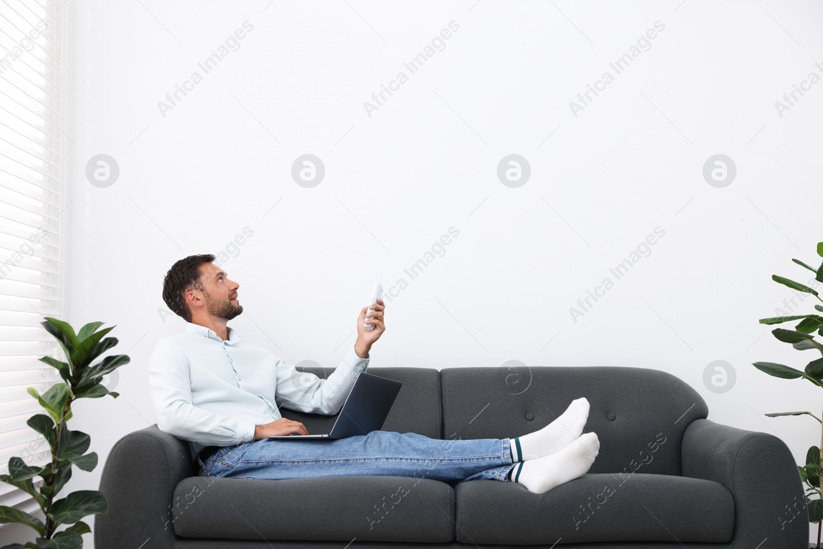Man using remote to control air conditioner on sofa at home. Space for text Photo of Man using remote to control air conditioner on sofa at home. Space for text