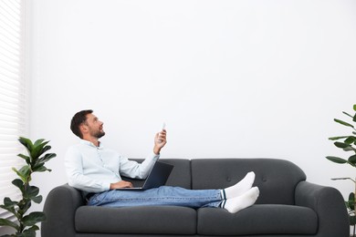 Photo of Man using remote to control air conditioner on sofa at home. Space for text