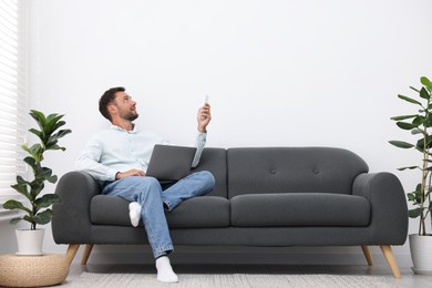 Photo of Man with laptop using remote to control air conditioner on sofa at home. Space for text