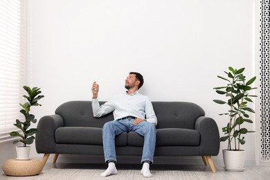 Photo of Man using remote to control air conditioner on sofa at home