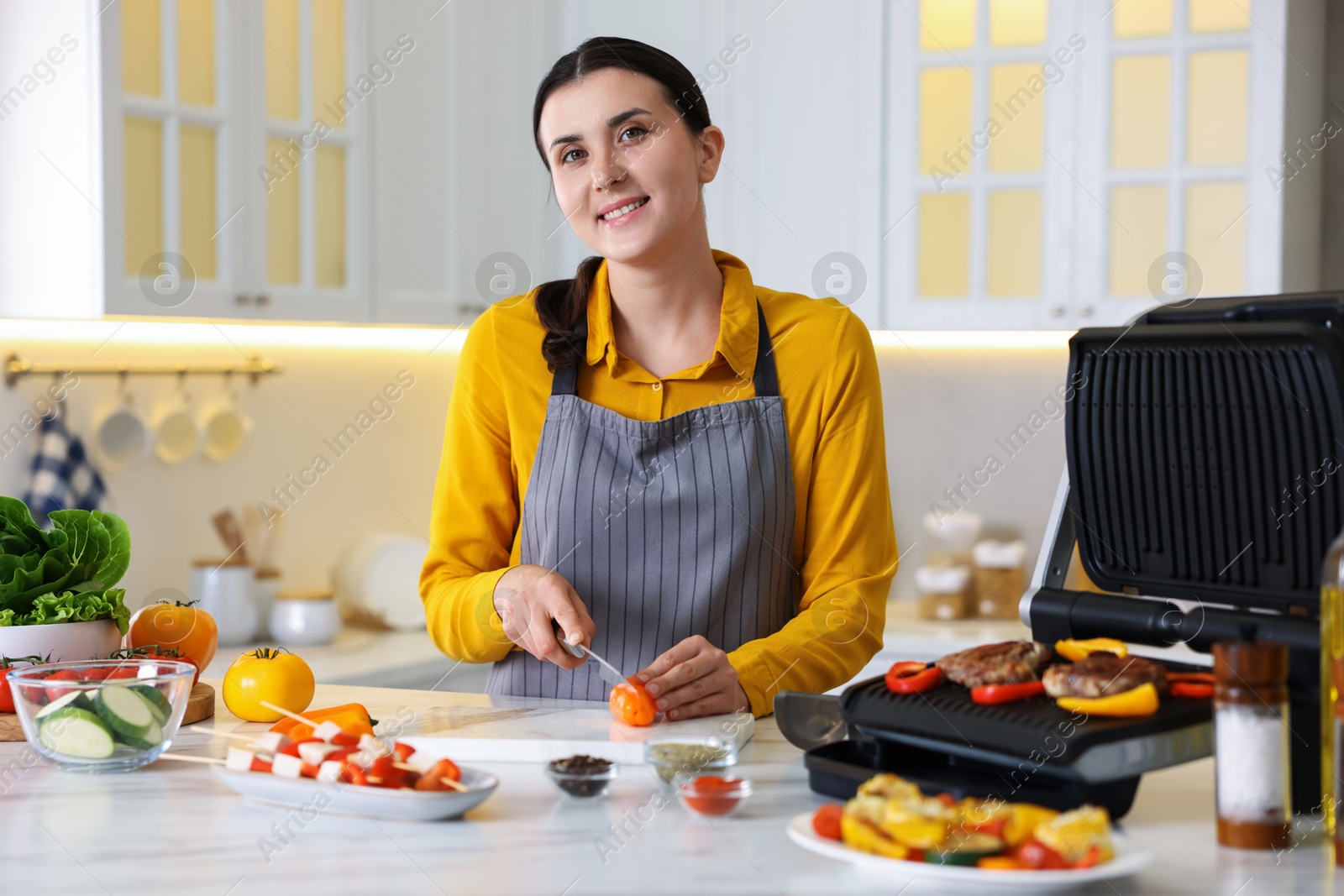 Photo of Woman cutting pepper while cooking meat and vegetables on electric grill in kitchen